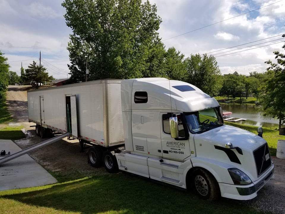 A white semi-truck with an attached trailer parked on a gravel surface near a grassy lawn and a small body of water.
