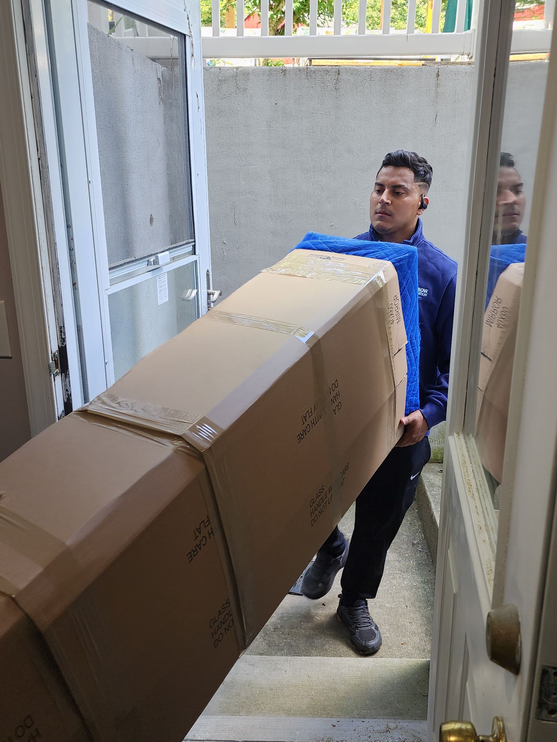 A person carries a large cardboard box and blue item up steps into a doorway.