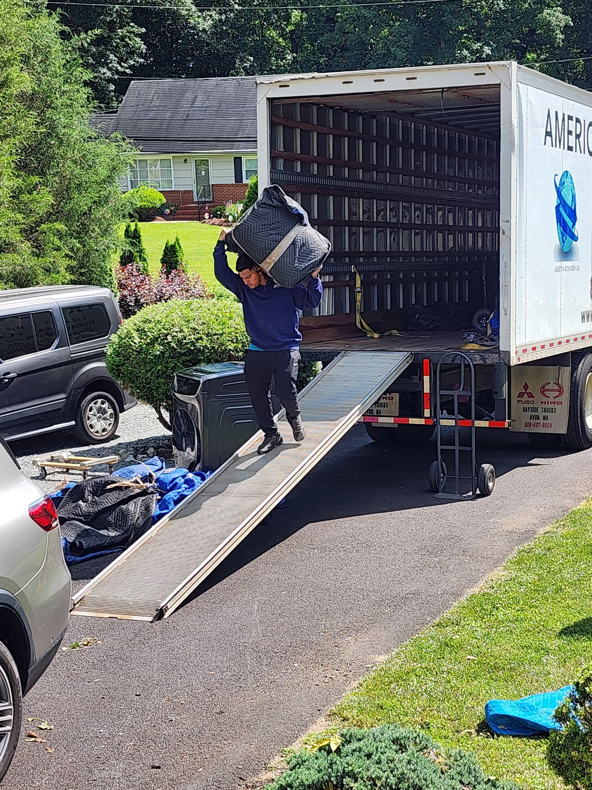 A person carries a black container down a ramp from a moving truck parked in a driveway.