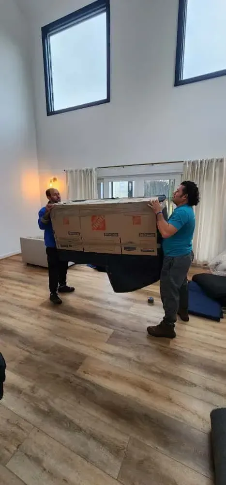Two movers carrying a large, wrapped object down a hallway; a third person is partially visible.