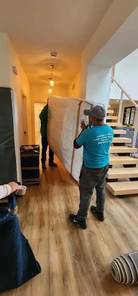 Movers carrying wrapped furniture in a living room with stairs and a window in the background.