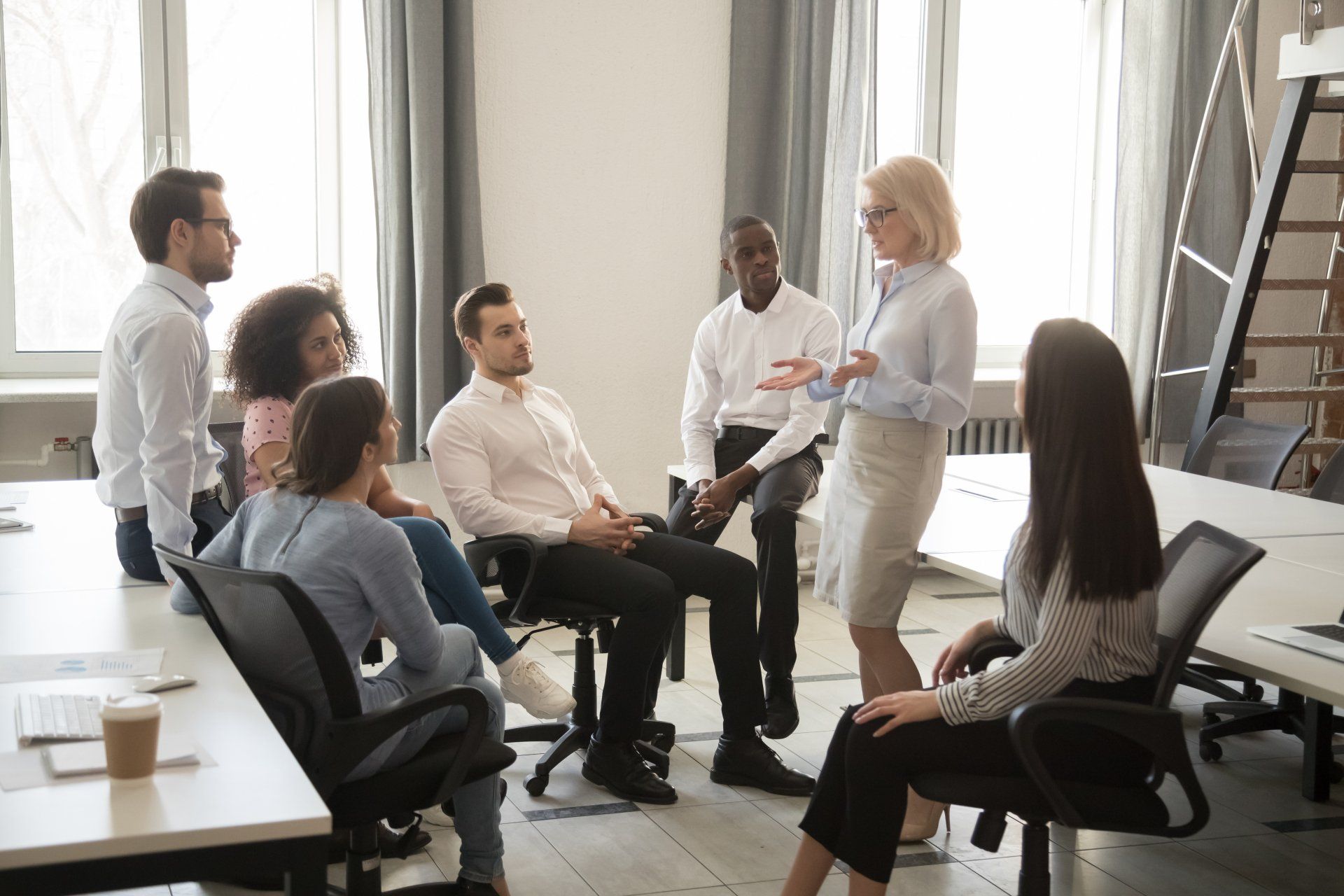 The vast majority of employee struggle with impostor syndrome at one time or another. You can fight it. Seven employees sitting and standing in a meeting room, one speaking to the others.