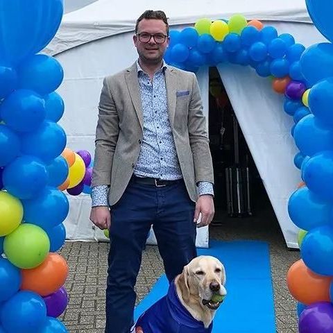 Man in blazer en hond in regenjas poseren bij een ballonnenboog.