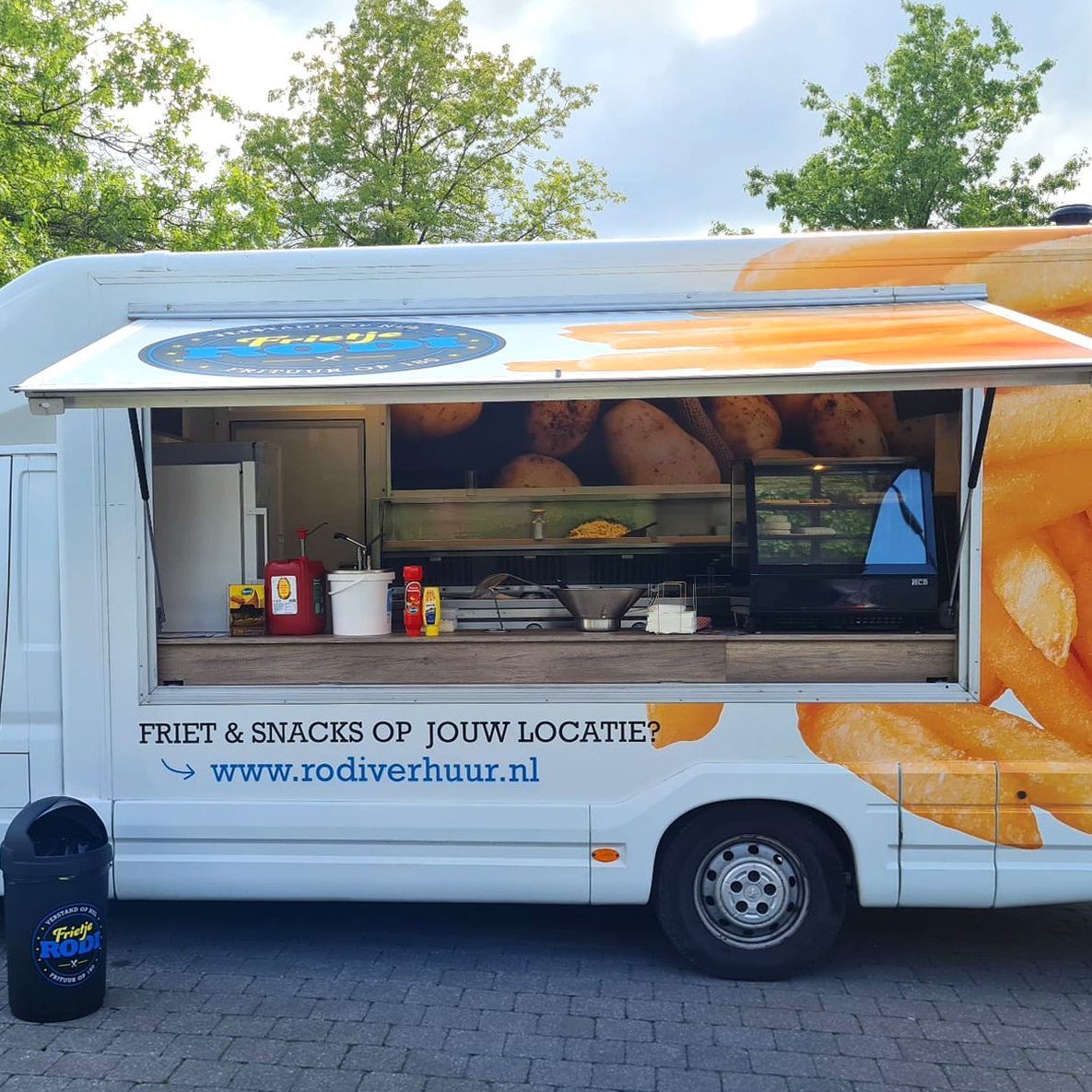 Foodtruck met open raam, friet en een bereidingsplek. Witte buitenkant met tekst en logo, buiten geparkeerd.
