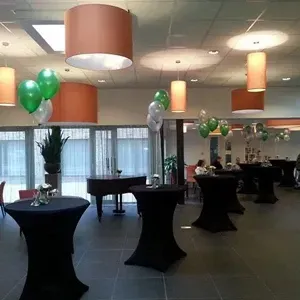Evenementenruimte ingericht met zwarte tafels, ballonnen en oranje/witte lampen; een piano op de achtergrond.