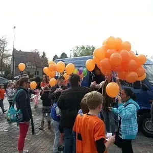 Mensen, waaronder kinderen, houden oranje ballonnen vast bij een busje en gebouwen.