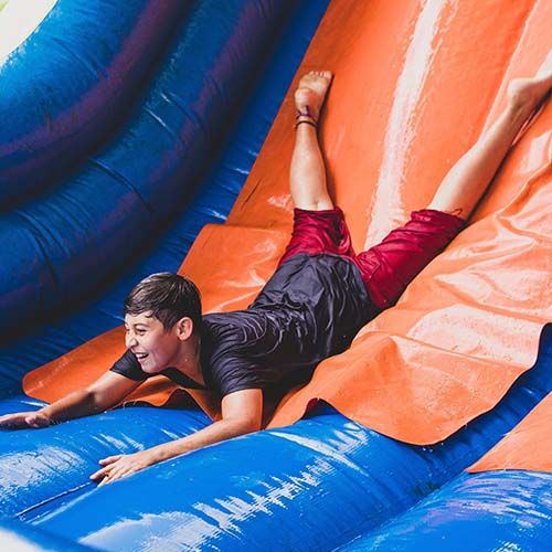 Jongen glijdt van een waterglijbaan, benen gespreid, lachend. Oranje en blauwe opblaasbare glijbaan.