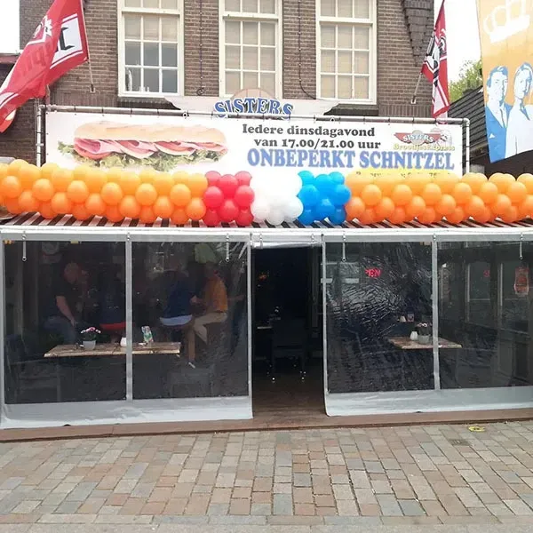 Restaurantgevel met oranje-rood-wit-blauwe ballonnenboog. Bord: 