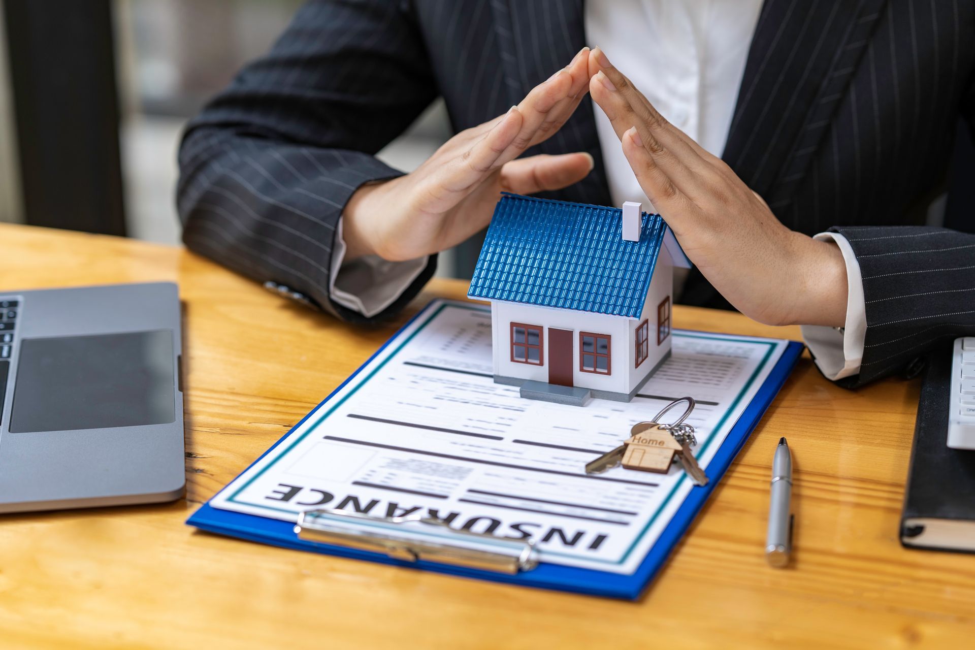 Person's hands protecting a miniature house on an insurance document, keys, and a pen on a desk.