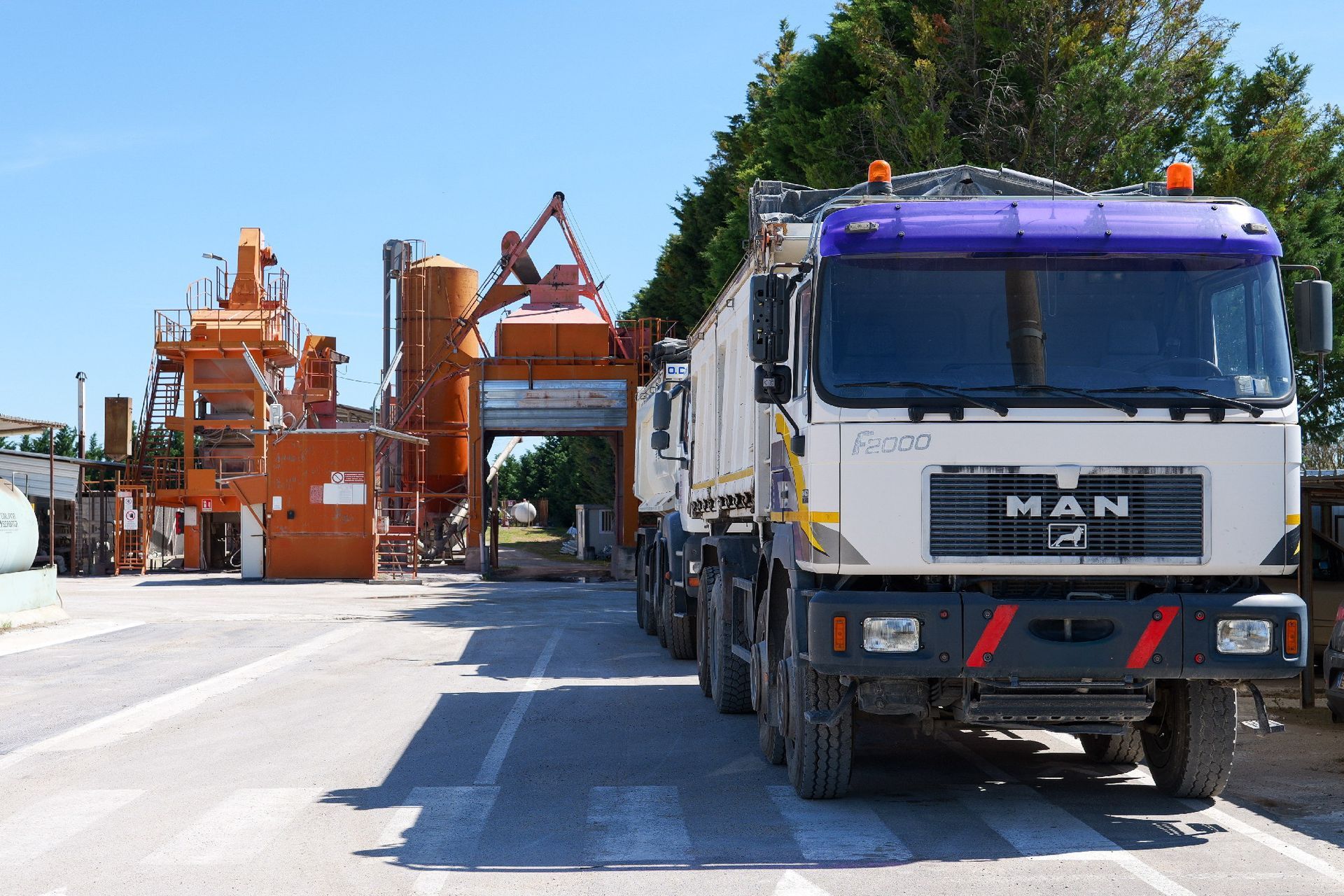 Un camion bianco accanto a macchinari industriali arancioni su una strada soleggiata con alberi sullo sfondo.