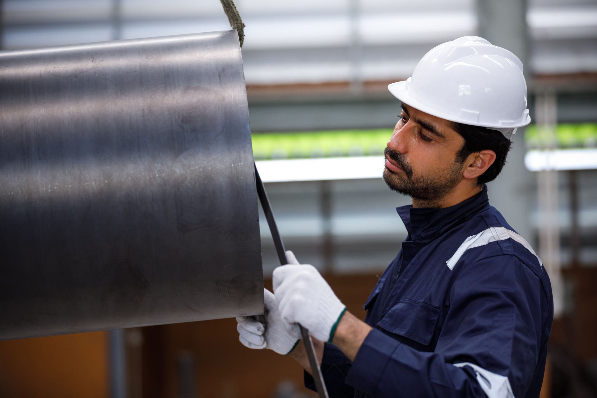 A worker is measuring a large hanging metal pipe using a ruler in a factory.