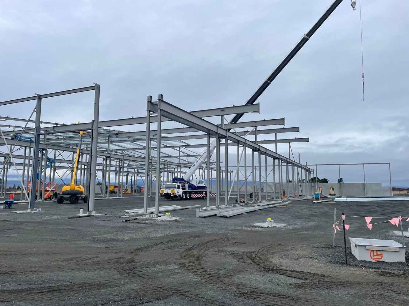 Construction site with steel framework for a building. Cranes, machinery, and workers are present under a cloudy sky.