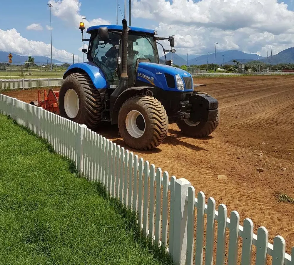 Blue Tractor Working on a Dirt Track Beside a White Picket Fence Under a Cloudy Sky — Tropical Lawns in Mulgrave, QLD