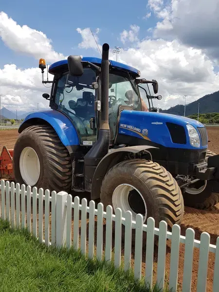 Blue Tractor Parked Beside a White Picket Fence Under a Cloudy Sky — Tropical Lawns in Mulgrave, QLD