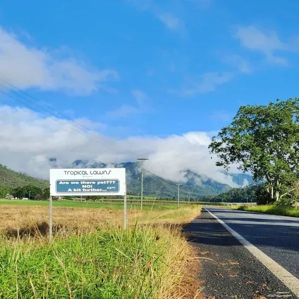 Road Through Tropical Valley With a Roadside Sign, Green Fields, Mountains, and Blue Sky — Tropical Lawns in Mulgrave, QLD