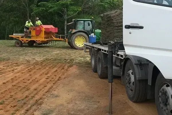 White Truck Towing a Red Farm Machine Along a Muddy Dirt Road in a Wooded Area — Tropical Lawns in Mulgrave, QLD