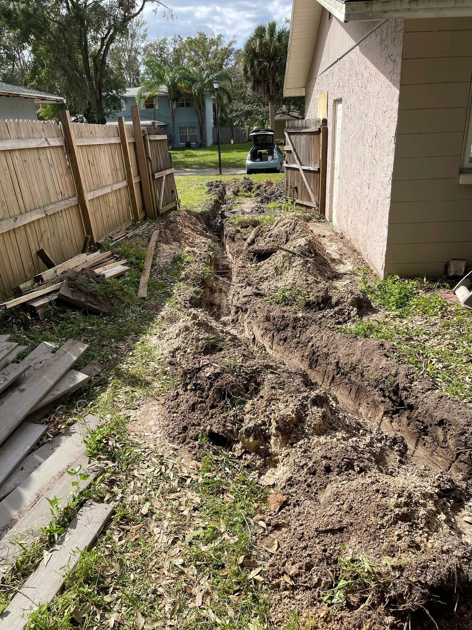 A narrow dirt trench in a grassy backyard, alongside a building and fence.