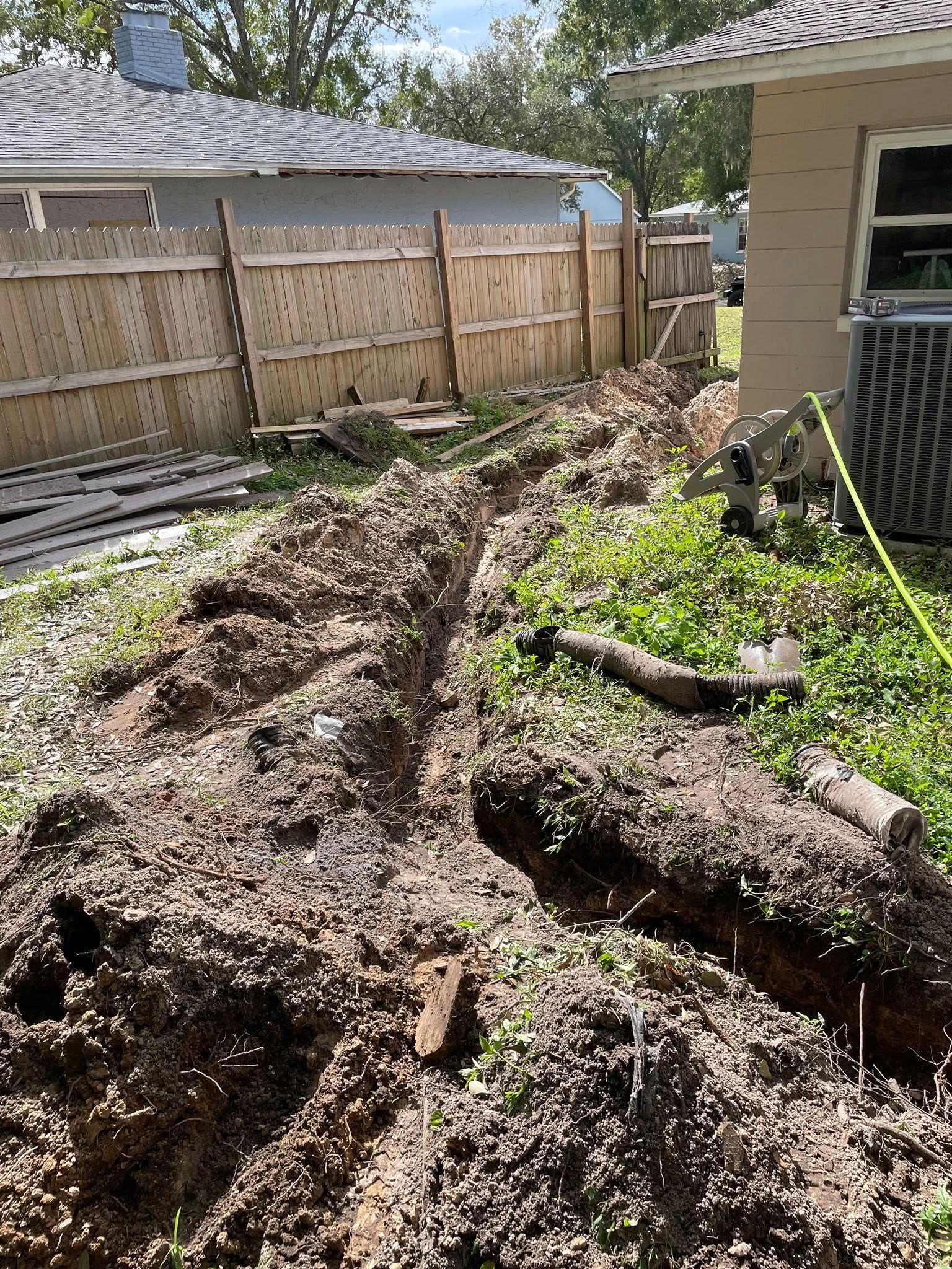 A trench dug in a grassy backyard, running parallel to a wooden fence and a house.