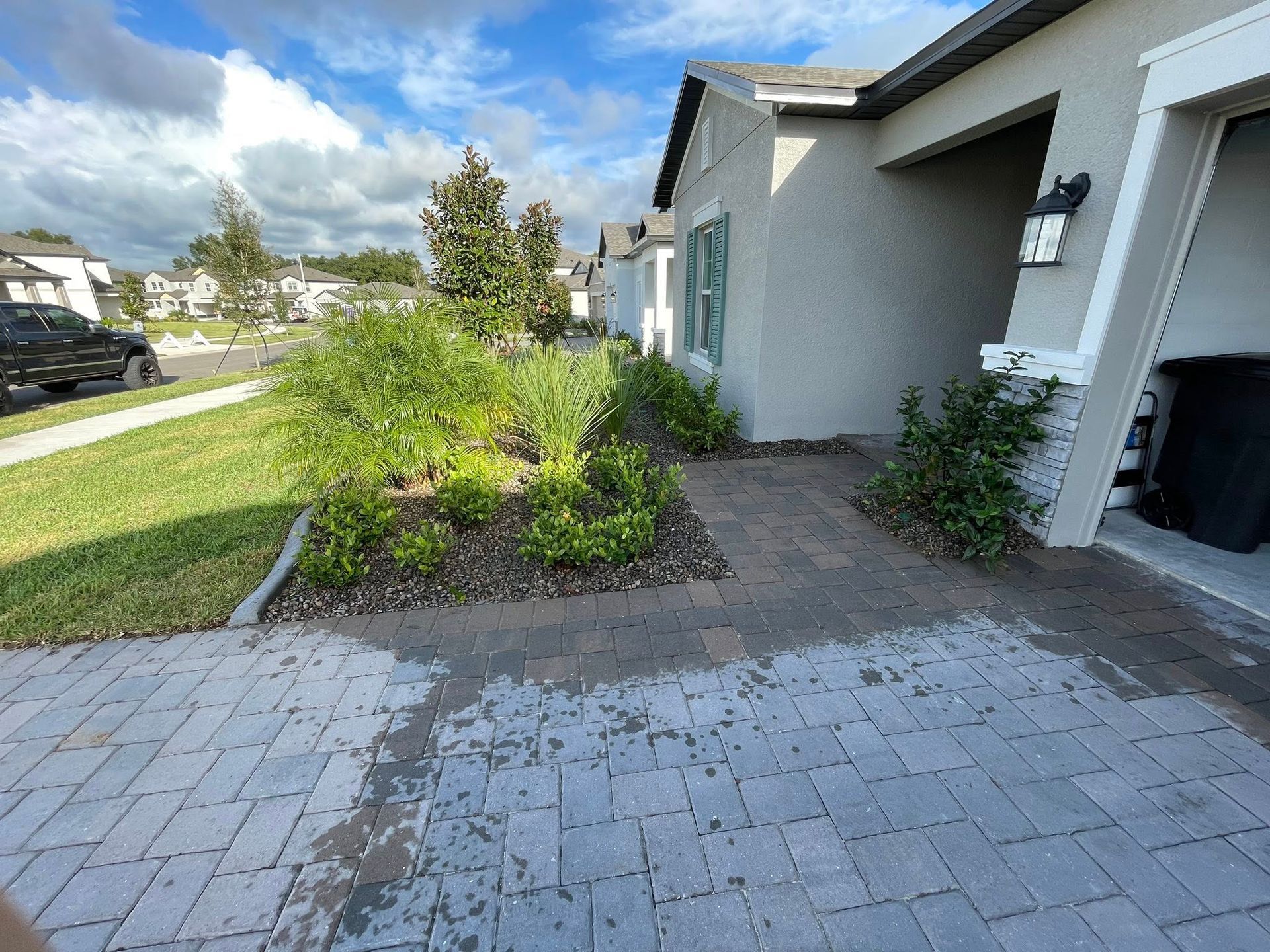A house exterior with brick driveway and garden beds. Green plants and gray siding. Bright sunny day.