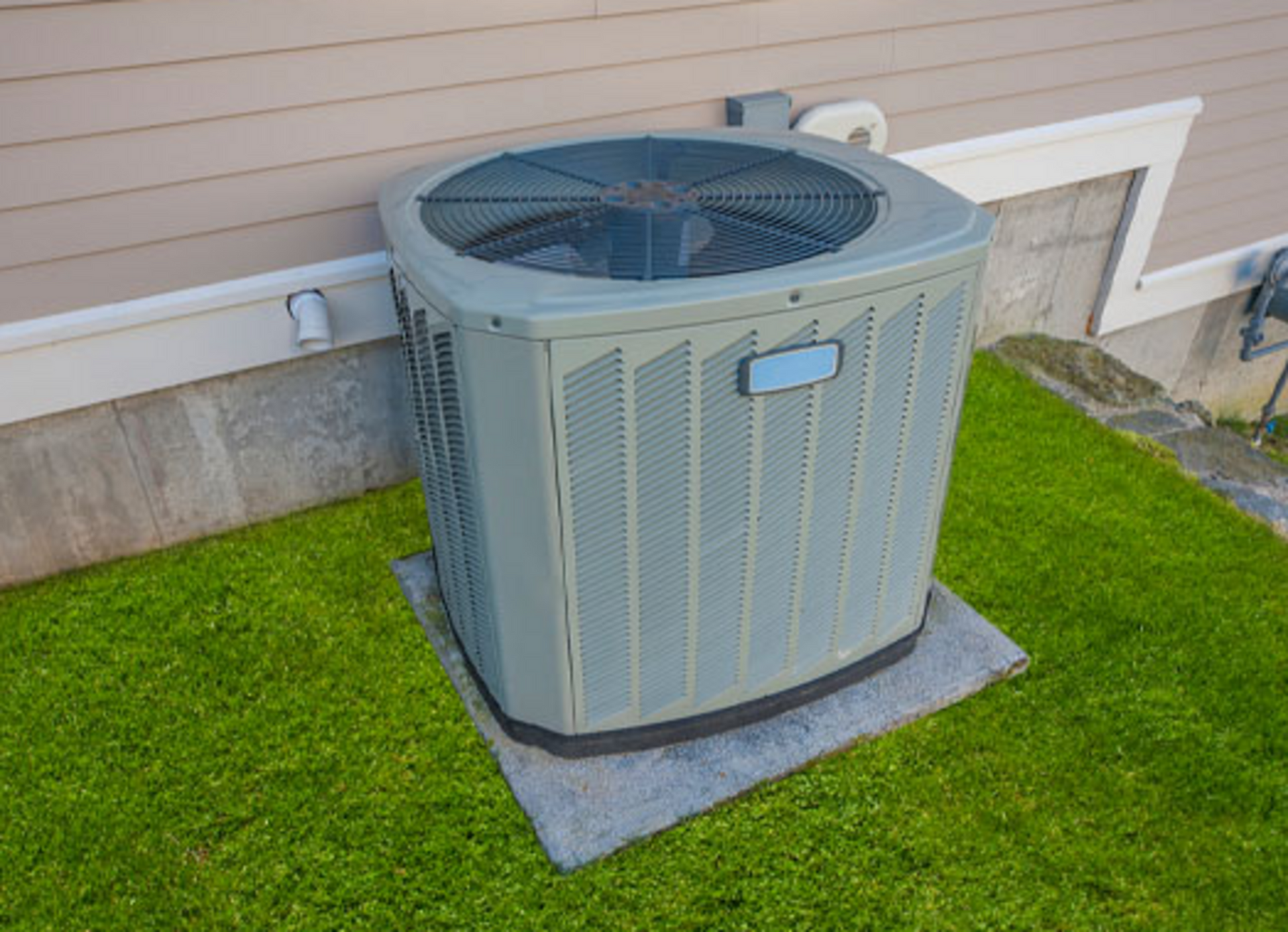 Gray air conditioning unit on a concrete pad, on a grassy lawn next to a beige house.