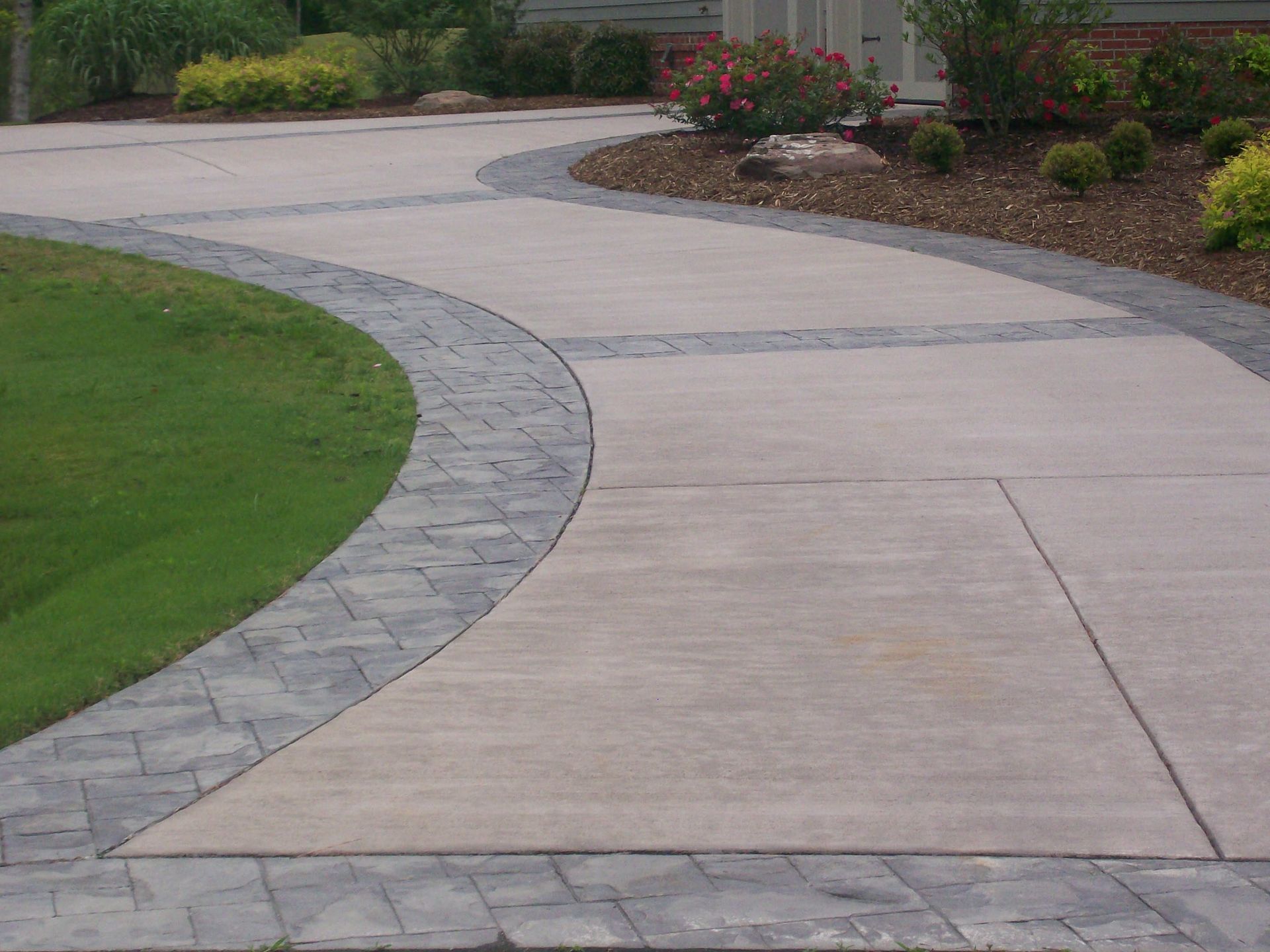 Curved concrete driveway bordered by stone pavers, green lawn, and landscaped flowerbeds