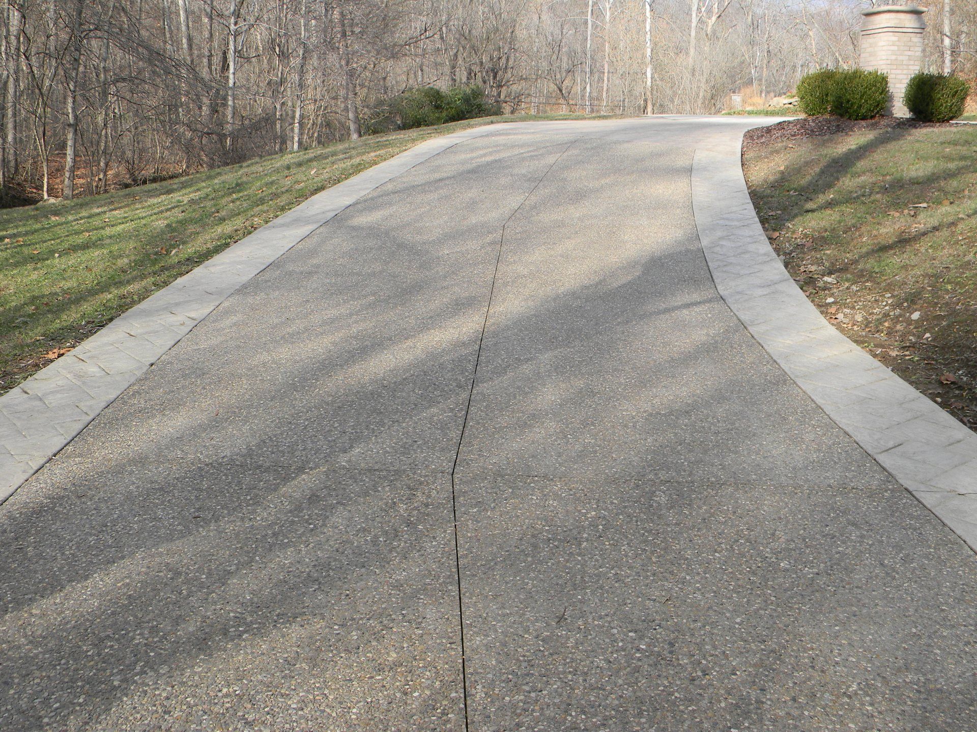 Curved paved driveway bordered by grass and trees, leading uphill toward a house in the distance