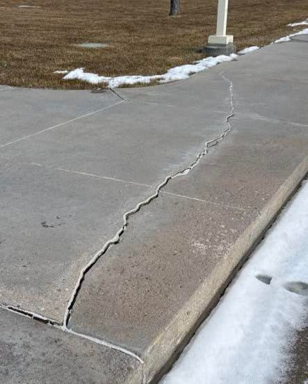 Cracked concrete sidewalk with a raised, uneven seam beside a curb and patchy snow