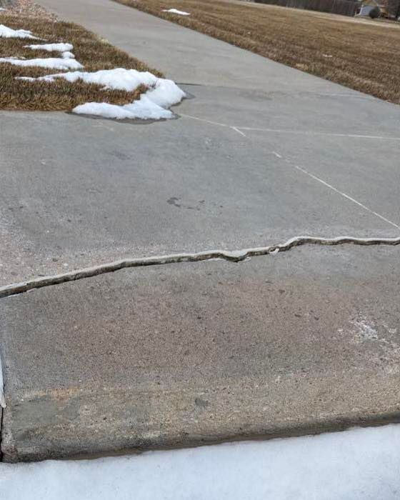 Cracked concrete sidewalk with snow along the edges and a patch of brown grass nearby