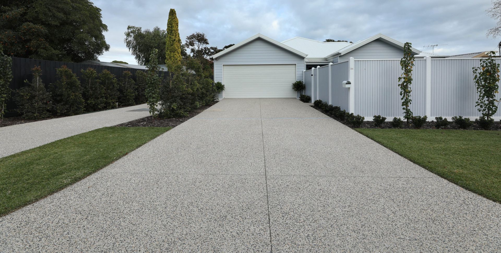 Long gravel driveway leading to a white garage, bordered by lawn, trees, and a white fence