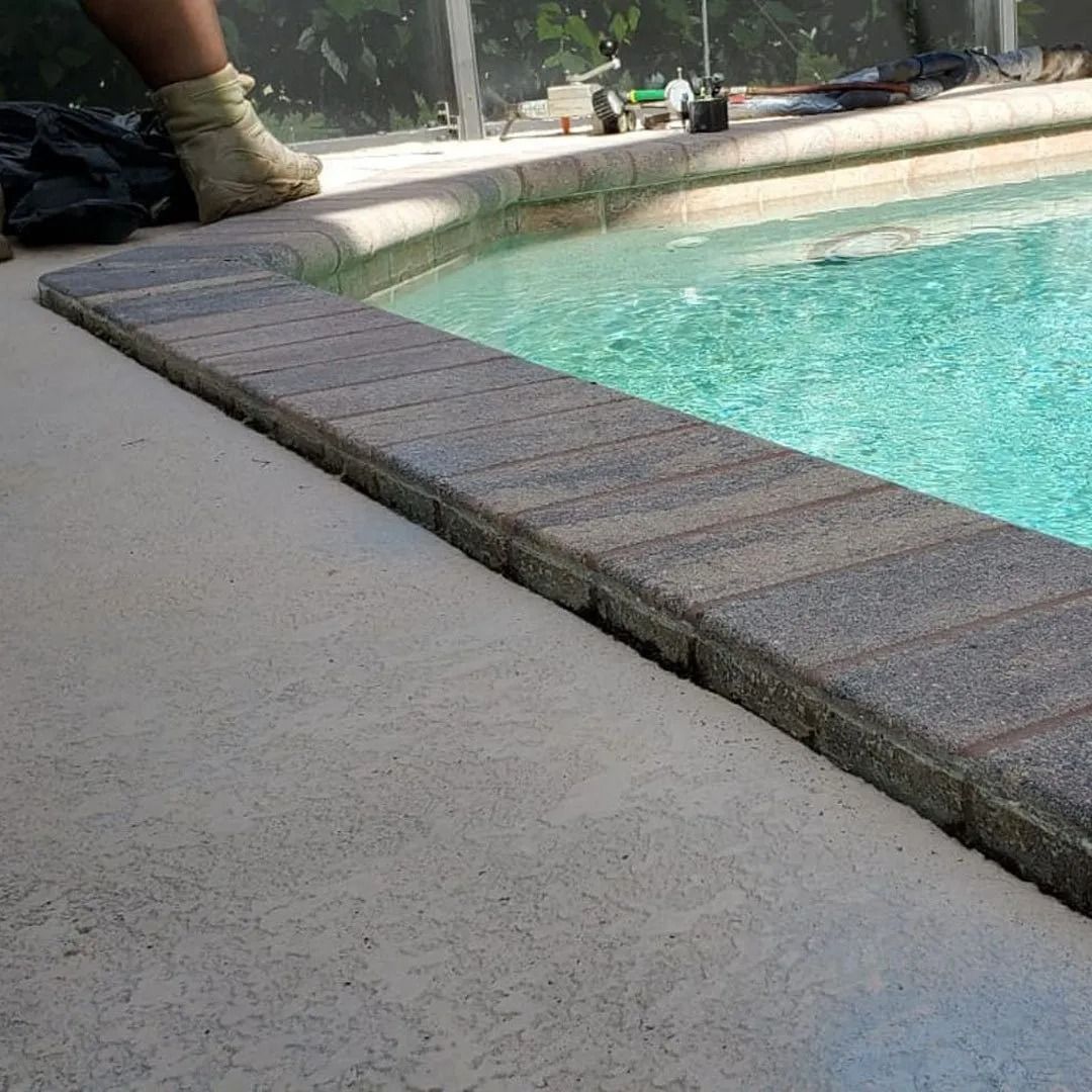 Pool edge with textured coping beside turquoise water; a person’s shoe is partly visible at left.