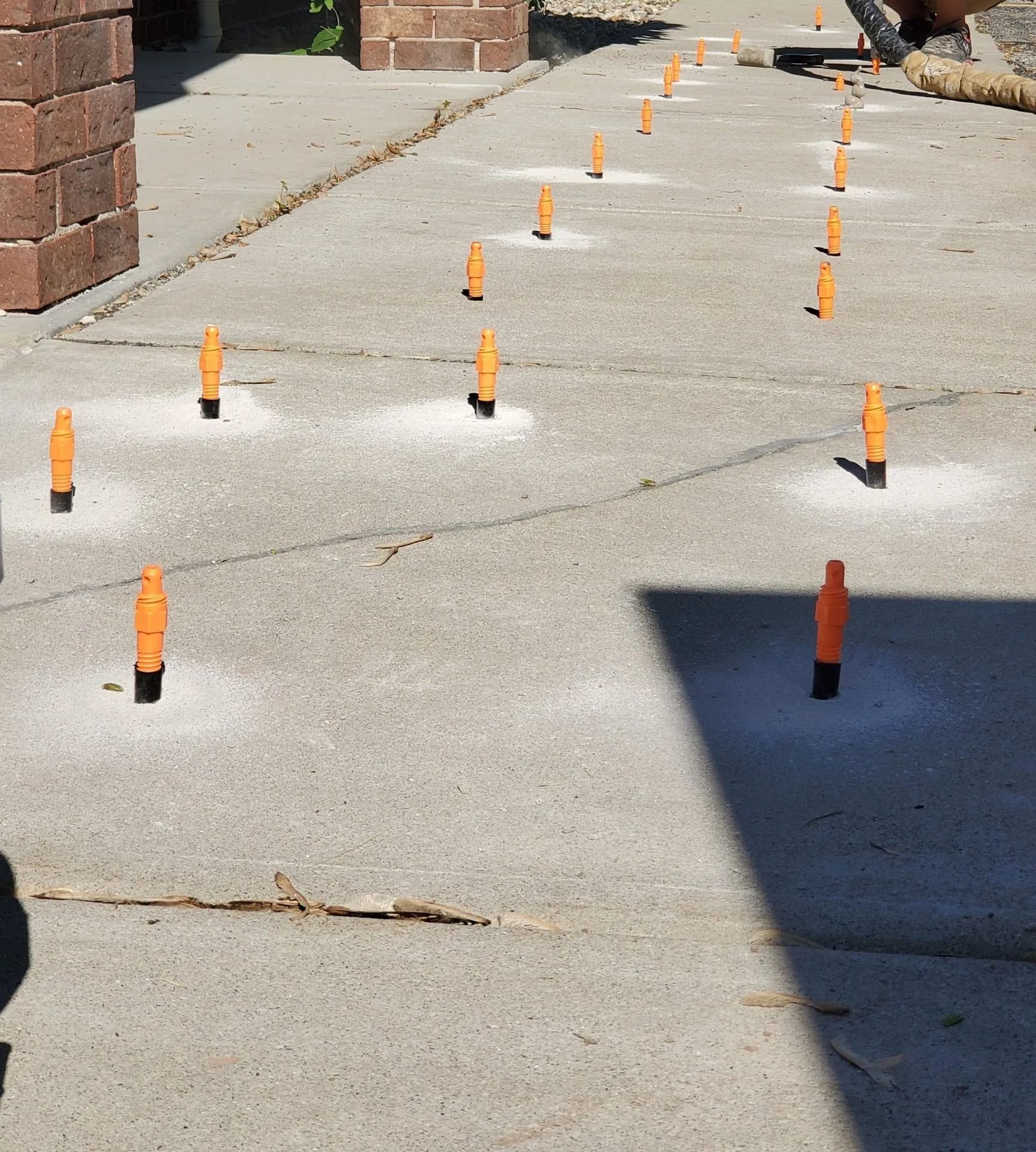 Orange utility markers lining a concrete sidewalk beside brick buildings, with sun and shadow.