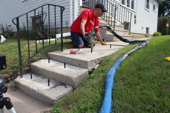 Person kneeling on front steps using a hammer, with blue tarp-lined grass beside the house