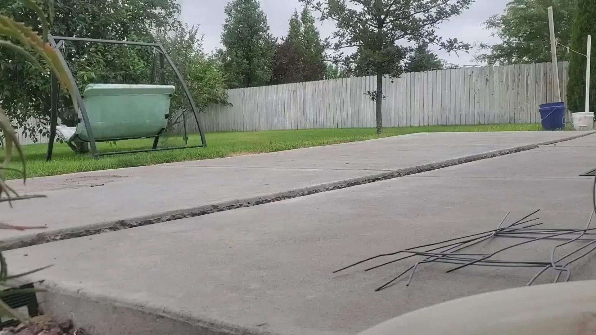 Backyard patio with a concrete slab, lawn, trees, wooden fence, and a green yard cart.