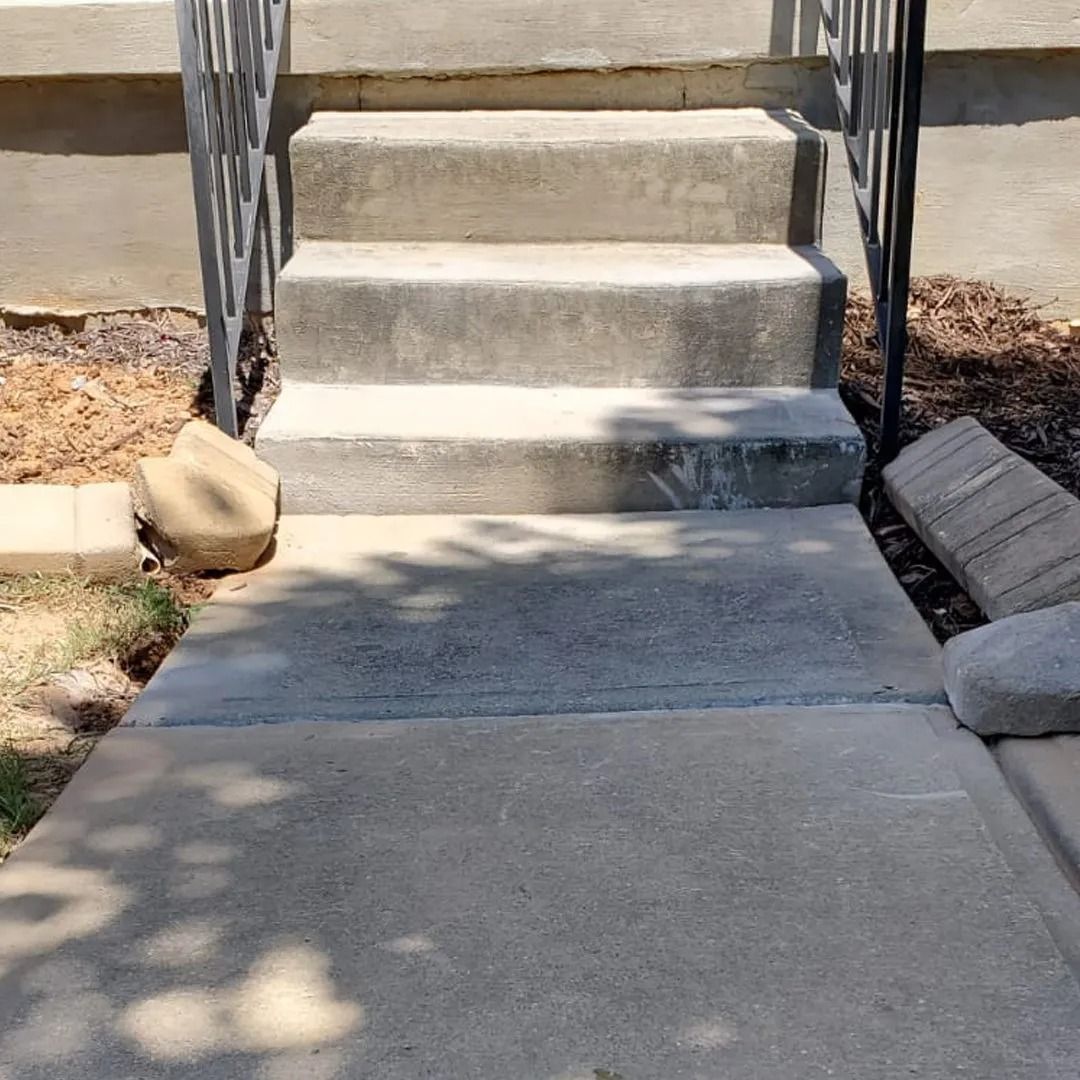 Concrete sidewalk leading to three concrete steps with black railings and stone borders