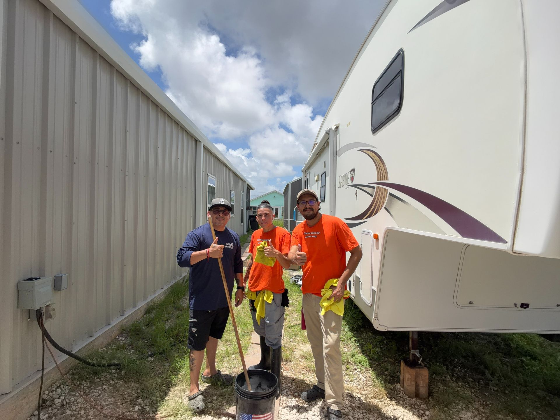 Three workers standing beside a large RV outside a metal building, one holding a bucket of tools.