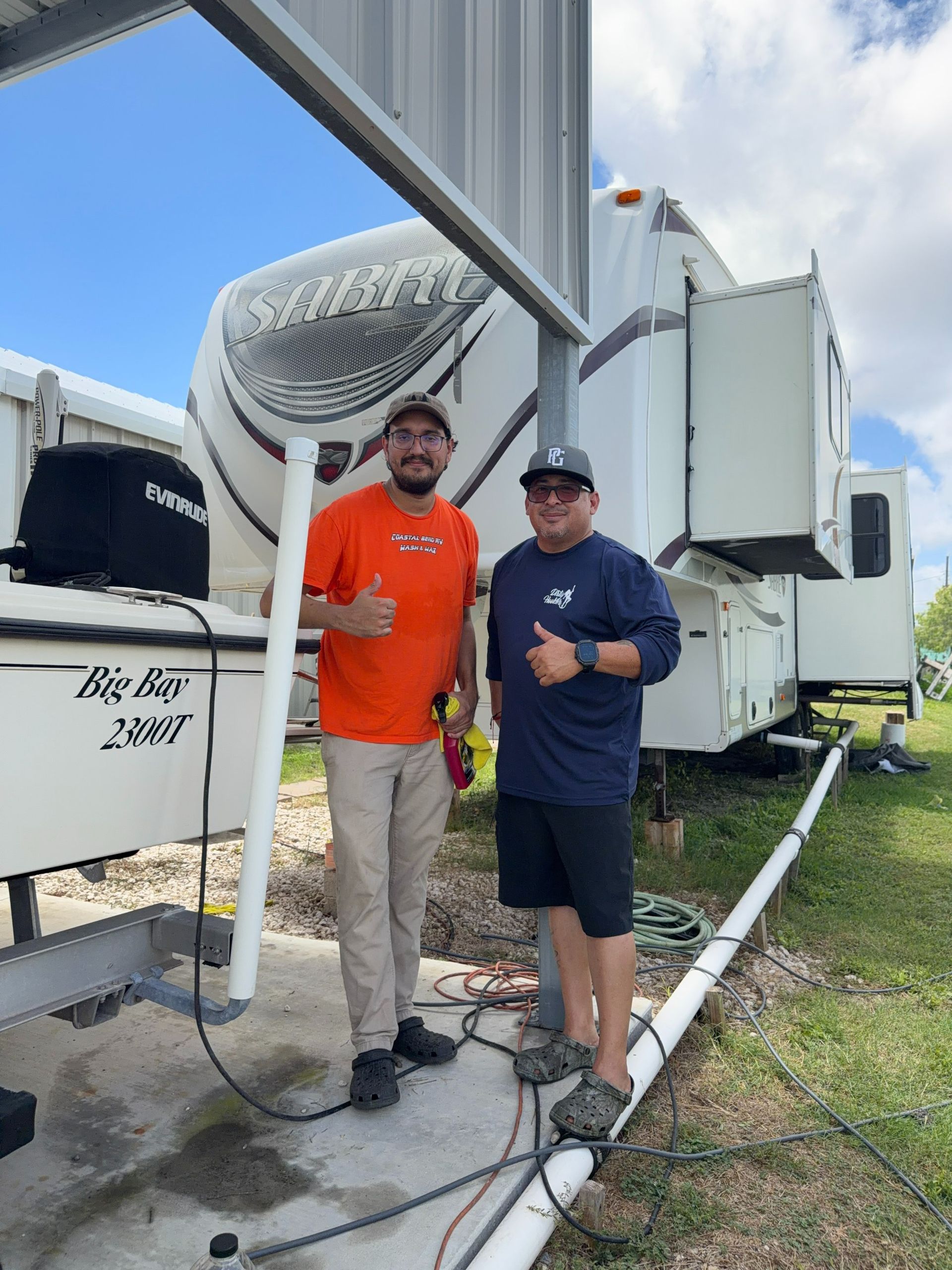 Two people posing beside a large RV at a campsite, one giving a thumbs-up.