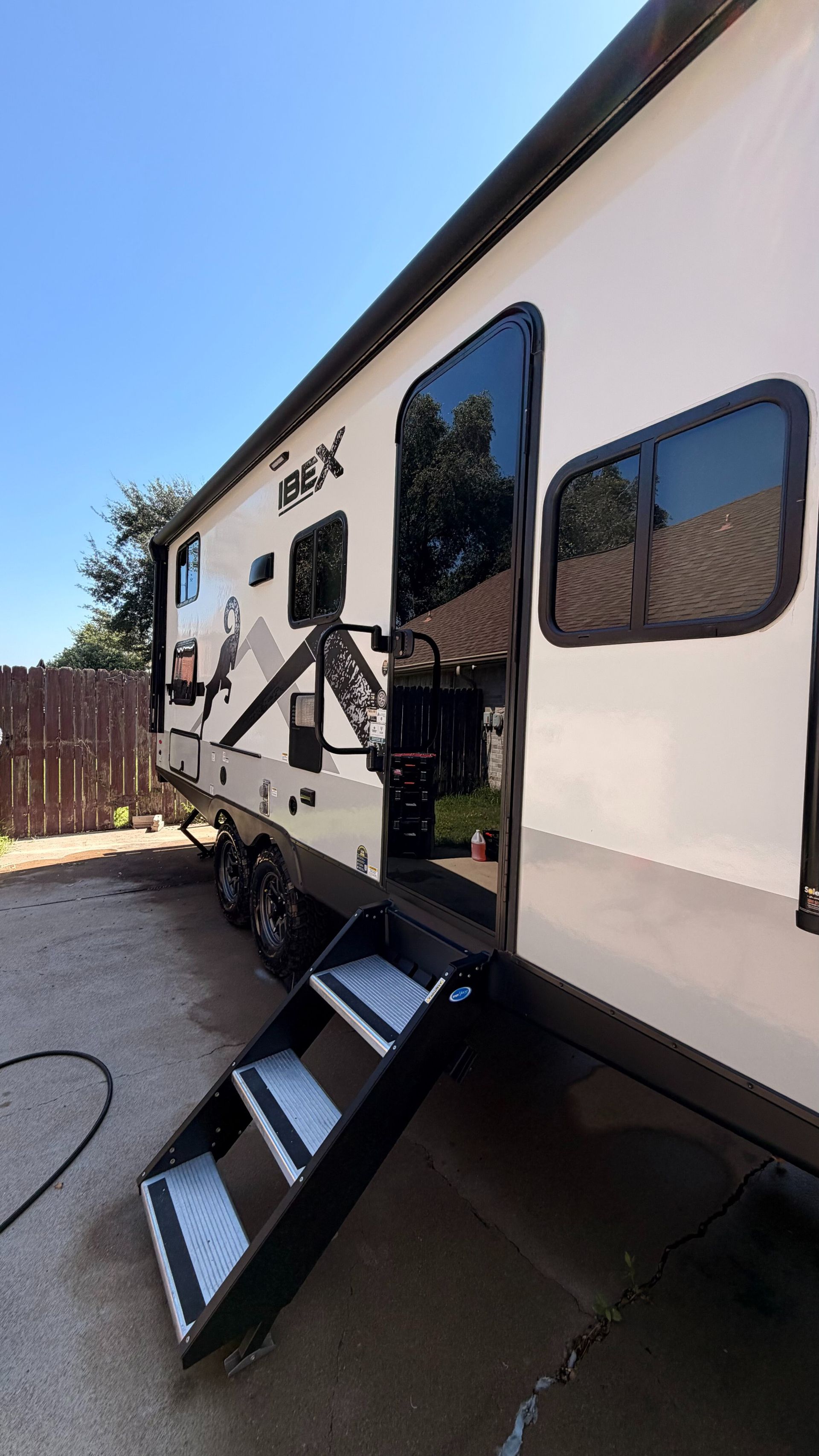White RV trailer with open door and steps parked beside a fence on a sunny driveway.