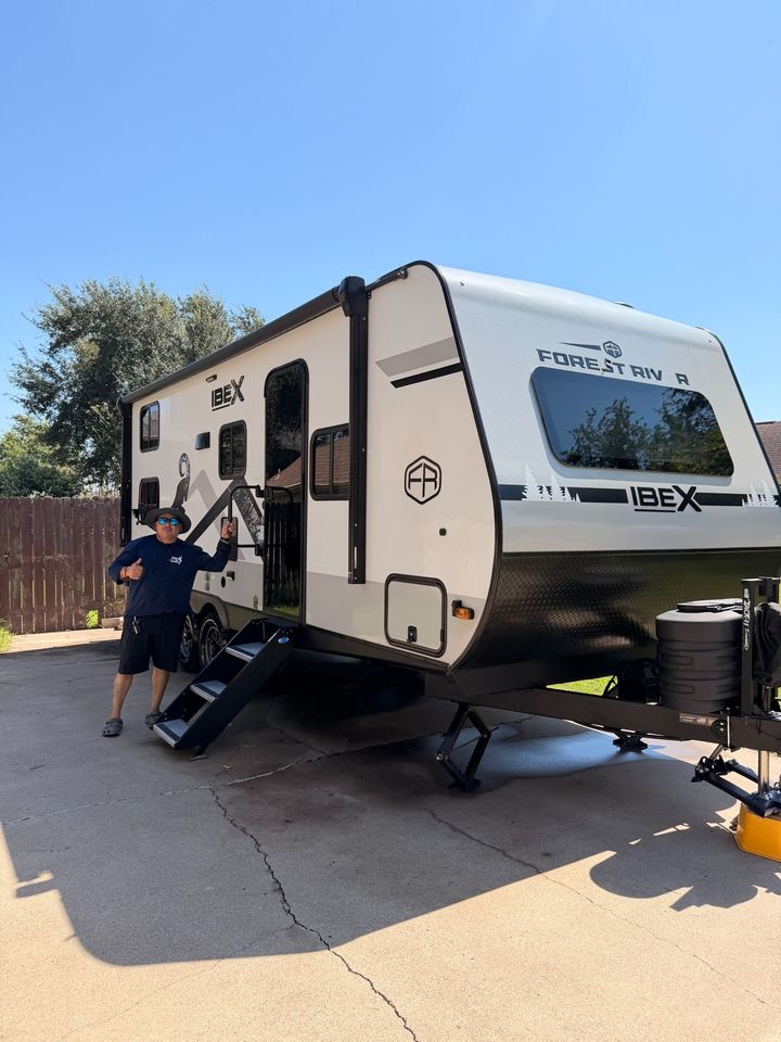 Person beside white travel trailer with black trim and extended awning on a sunny driveway