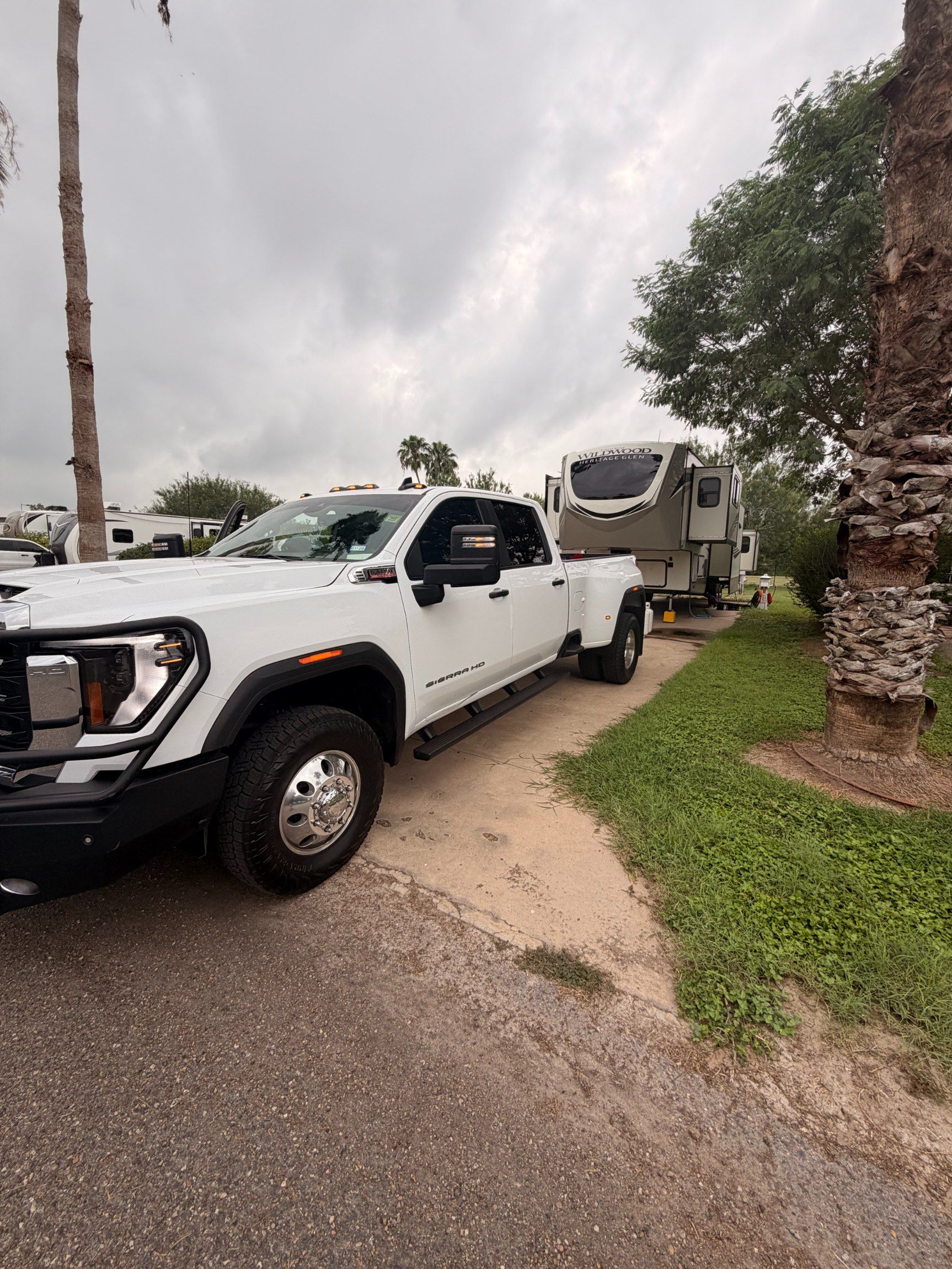 White pickup truck towing a trailer on a driveway beside a palm tree under cloudy skies