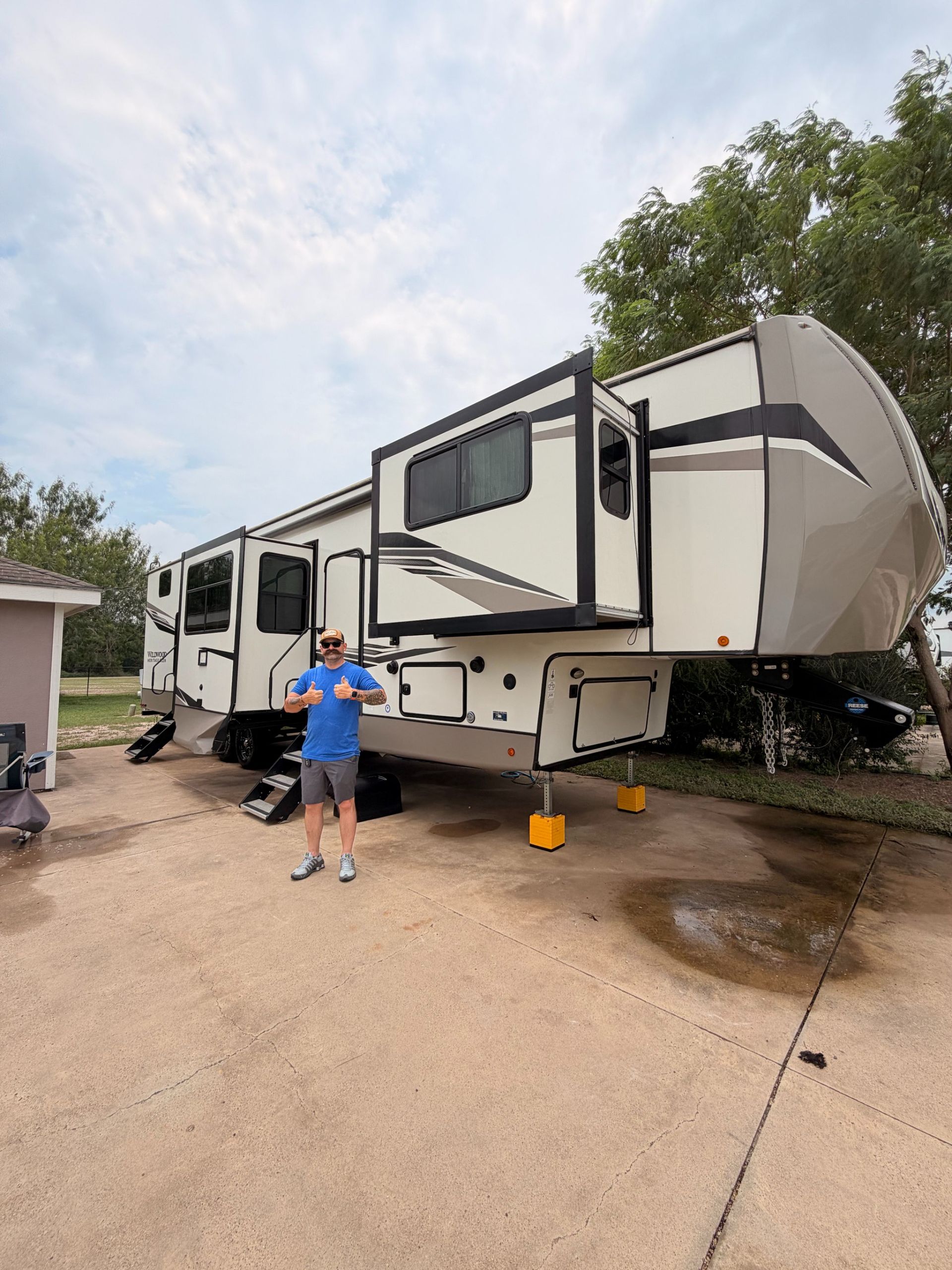 Large fifth-wheel RV with slide-outs parked on a wet driveway; person stands beside it.