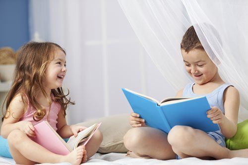 Two children, one in pink, one in blue, reading and smiling together under a sheer canopy.