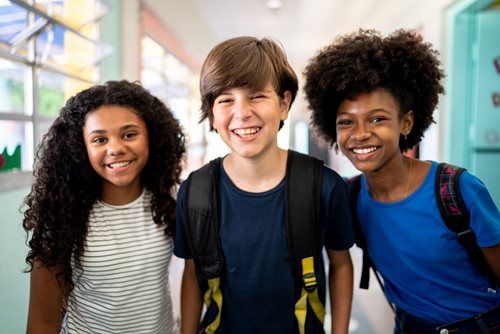 Three smiling students in a school hallway, wearing backpacks, looking at the camera.