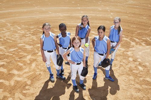 Five children in light blue shirts and backpacks stand on a sandy field, looking up at the camera.