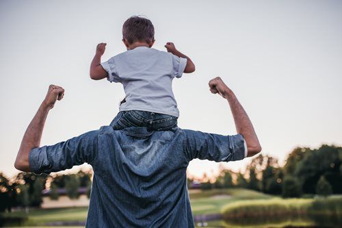 Father carrying child on shoulders, both with arms raised in the air, outdoors.