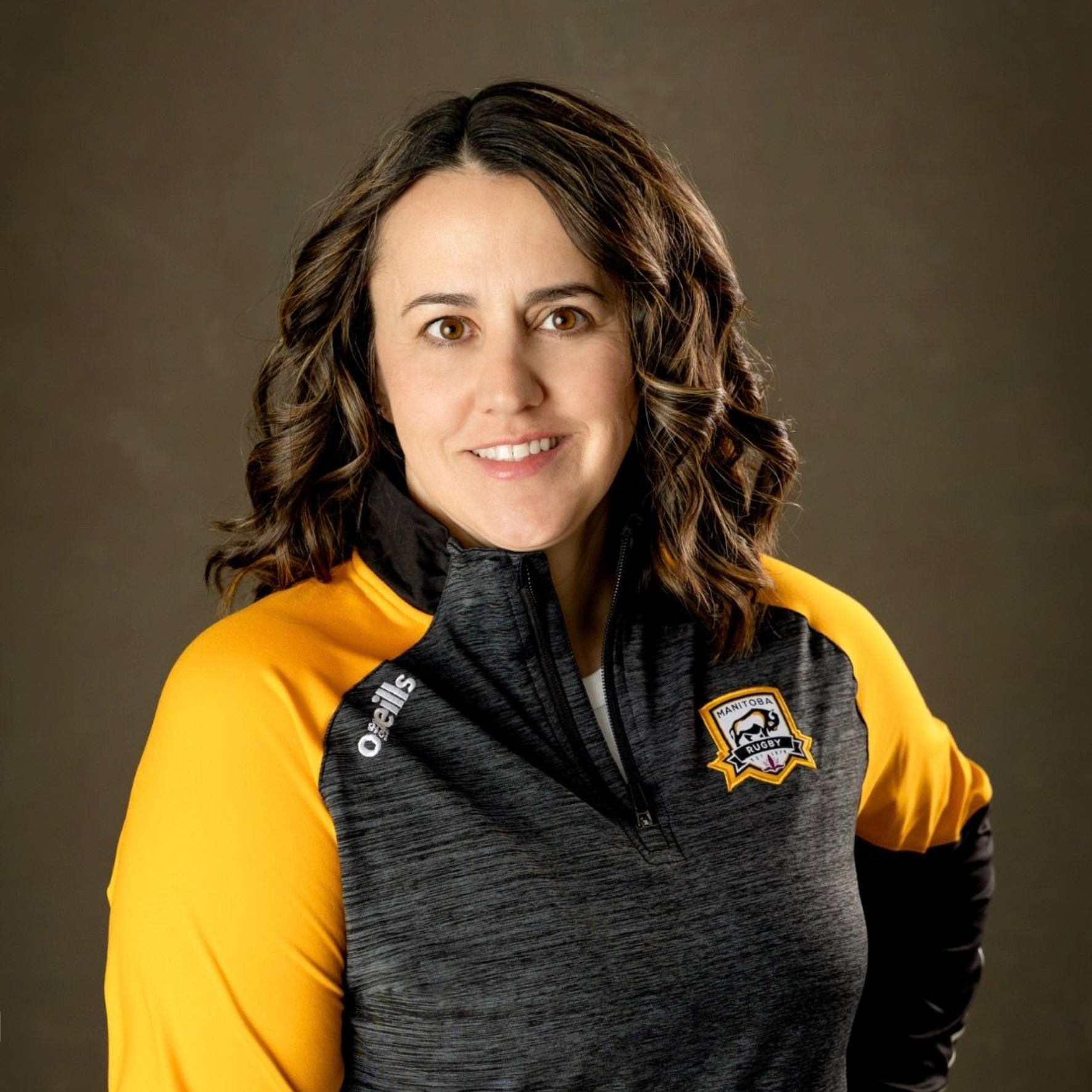 A smiling person wearing a black and yellow athletic zip-up jacket with a team logo, posing against a dark background.