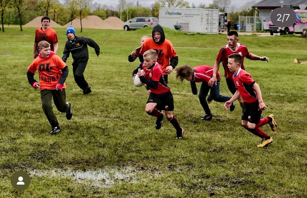 A group of people in red jerseys play rugby on a wet, muddy field. One person runs forward while carrying the ball.