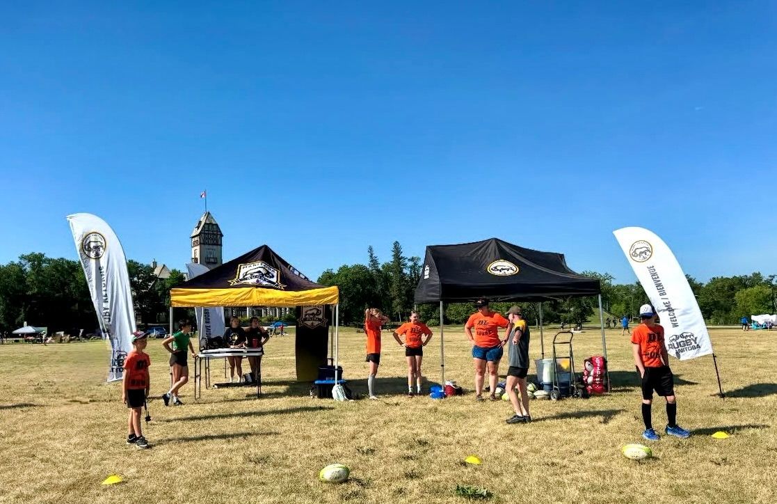 People in athletic wear stand near two event tents and banners on a sunny, grassy field.