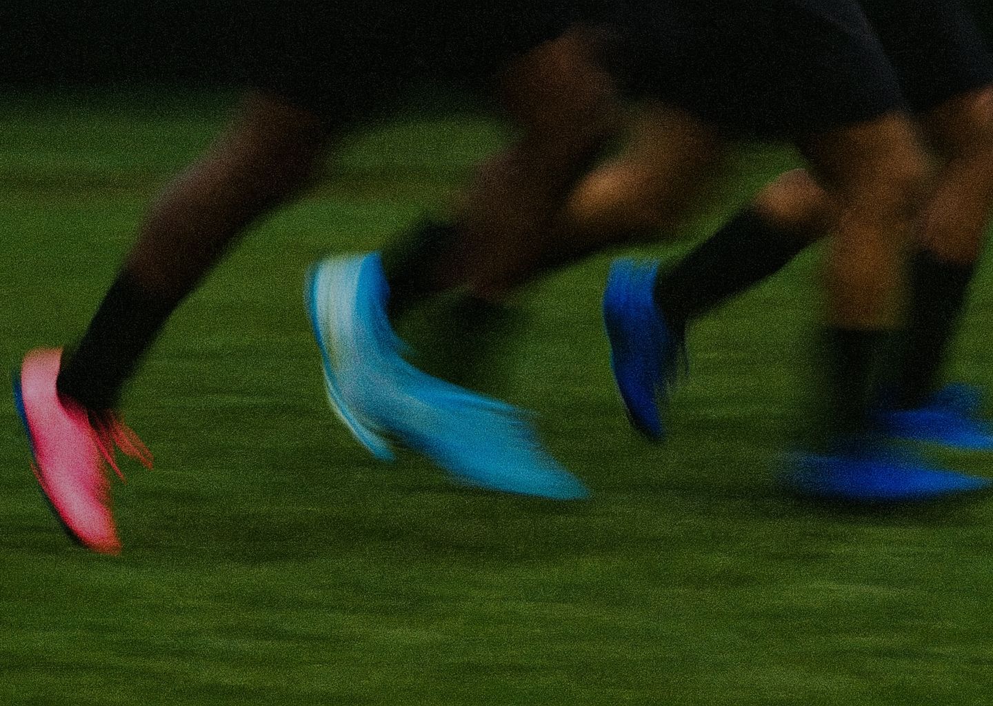 A close-up of rugby players' legs and feet in bright pink, blue, and light blue cleats running on a grassy field.