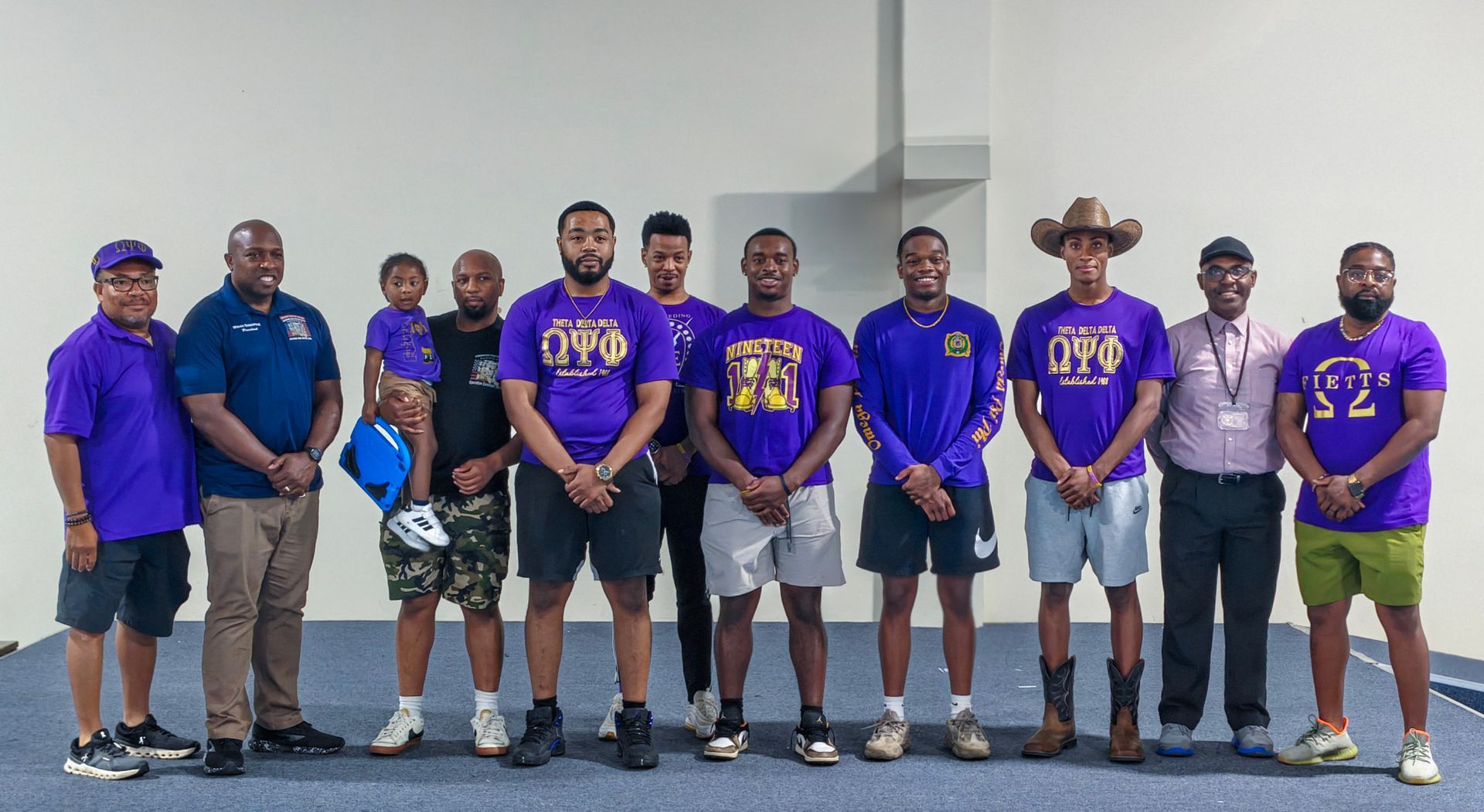 A group of people wearing purple shirts and casual clothing standing together in a line against a plain white wall.