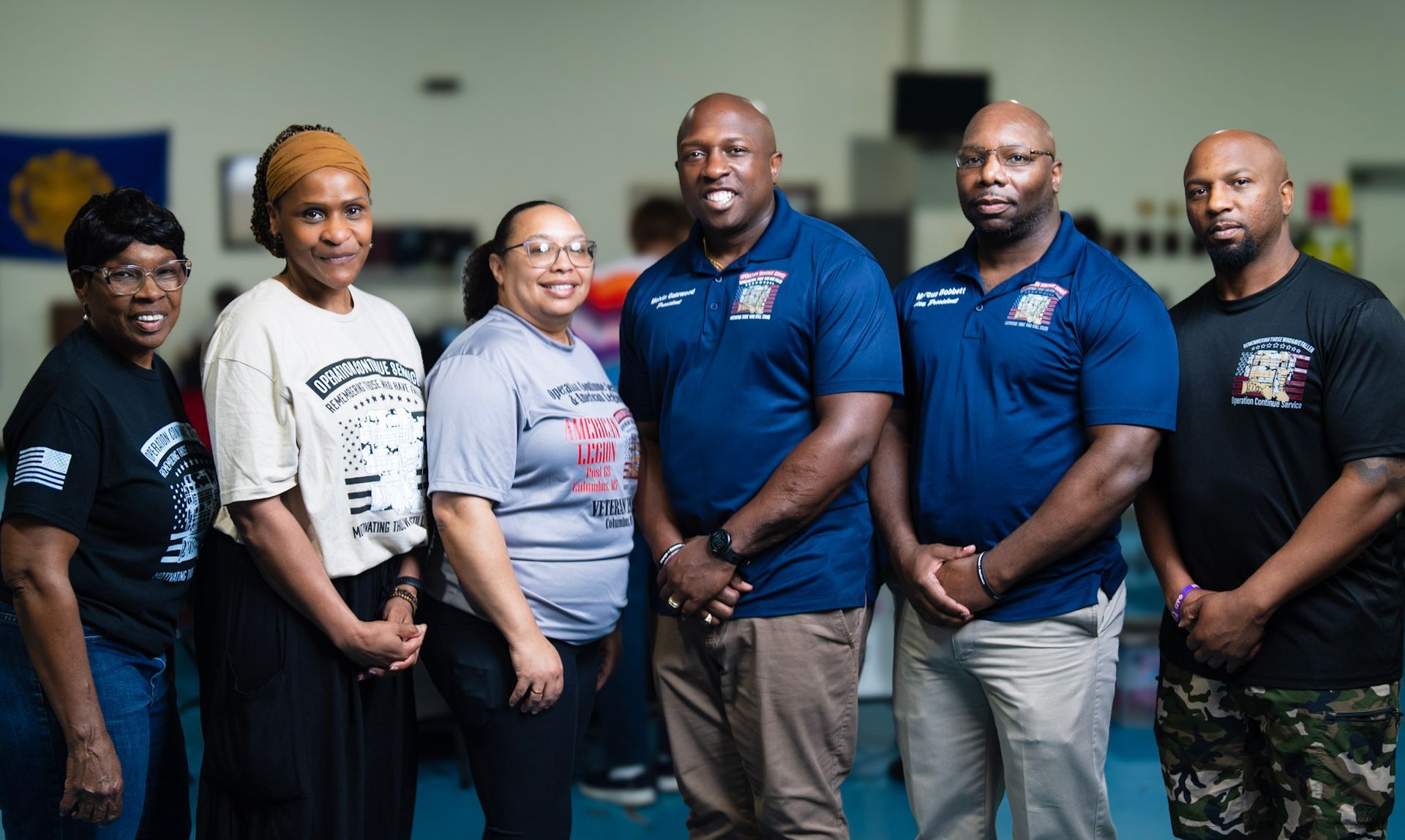 Six people stand side-by-side in a large room, smiling and posing for a group photo while wearing matching organization gear.